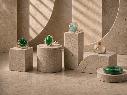 Luxury jade rings displayed on sculptural stone pedestals, featuring green jade, carved floral jade, pale blue jade, white jade, and gold details in a refined neutral gallery setting.